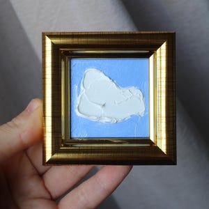 May include: A small, framed painting of a white abstract shape on a light blue background. The frame is gold.
