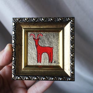Könnte beinhalten: Ein kleines quadratisches Gemälde einer roten Ziege mit gemusterten Hörnern vor einem strukturierten goldenen Hintergrund. Das Kunstwerk befindet sich in einem dunklen, verzierten Metallrahmen mit einem sich wiederholenden Bogenmuster am Rand.