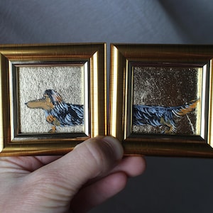 May include: Two miniature framed paintings of dachshund dogs. One shows the dog standing, the other shows the tail. The paintings have gold frames and gold leaf backgrounds. The dog's fur is black, brown, and tan.