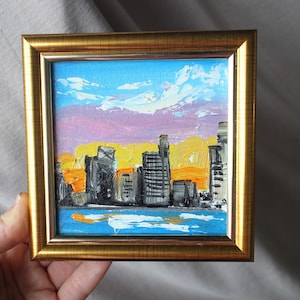 Puede incluir: Pequeña pintura al óleo enmarcada de un paisaje urbano al atardecer. La obra presenta un horizonte de edificios en tonos grises y negros, sobre un cielo de tonos azules, rosas y amarillos. La pintura está en un marco dorado.