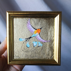 May include: A framed painting of a colourful bird with pink, blue and orange feathers flying against a gold background.