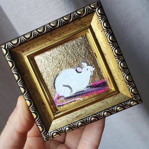 May include: A small framed painting of a white mouse on a gold background. The mouse is sitting on a pink and purple stripe.