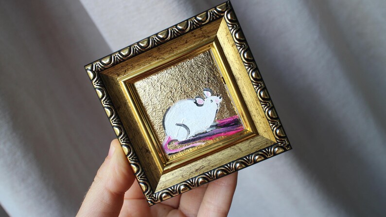 May include: A small framed painting of a white mouse on a gold background. The mouse is sitting on a pink and purple stripe.