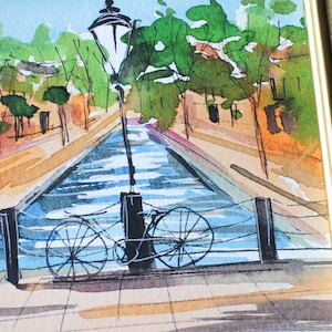 May include: A watercolour painting of a canal scene with a bicycle leaning against a lamppost. The canal is blue and the buildings on either side are painted in shades of brown and orange. The painting is framed in a gold frame.