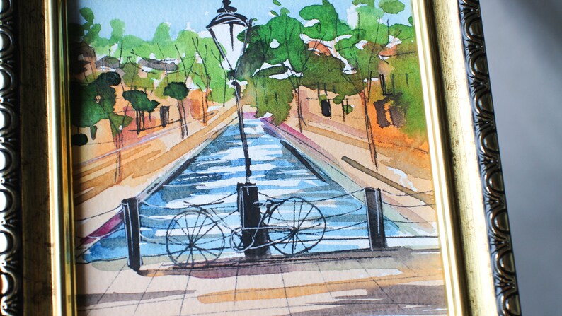May include: A watercolour painting of a canal scene with a bicycle leaning against a lamppost. The canal is blue and the buildings on either side are painted in shades of brown and orange. The painting is framed in a gold frame.