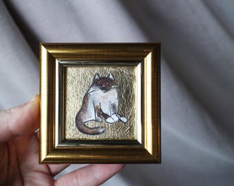 May include: A small, gold-framed picture of a white and brown cat sitting on a gold background.