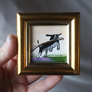 May include: A small, square painting in a gold frame depicts a black and white dog carrying a stick. The dog is painted with simple brushstrokes, set against a background of green grass and purple flowers.