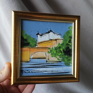 Small framed painting of a bridge and building with a gold frame. The artwork features a bridge with a yellow arch, a white building, and green trees against a blue sky and water.