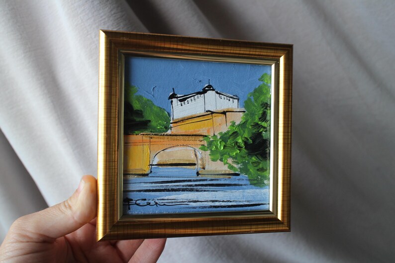 Small framed painting of a bridge and building with a gold frame. The artwork features a bridge with a yellow arch, a white building, and green trees against a blue sky and water.