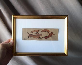 May include: A framed artwork featuring a circle of brown dachshunds on a tan background. The gold frame surrounds the artwork, which is set against a white mat. The artwork is signed in the lower left corner.