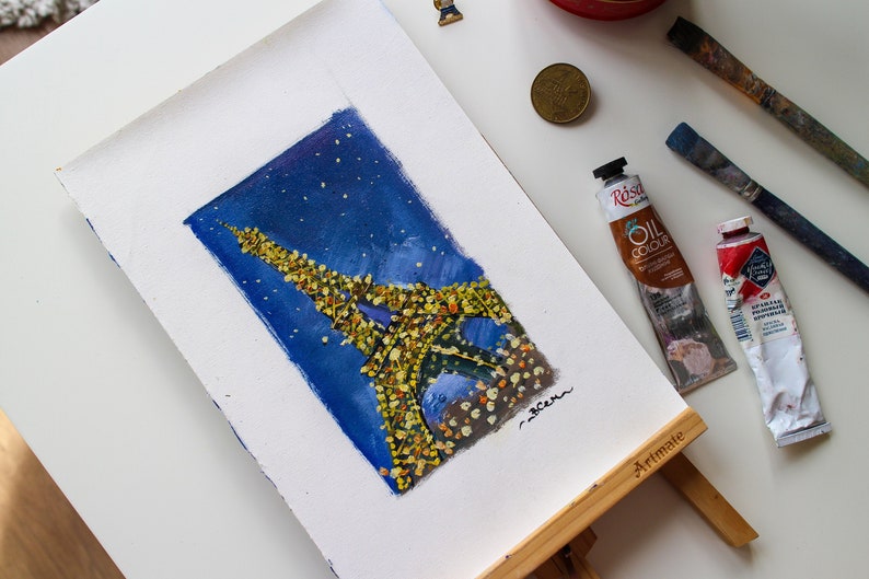 May include: A painting of the Eiffel Tower in Paris, France, with a starry night sky background. The tower is painted in yellow and white, with a blue sky and white stars. The painting is on a white canvas and is on an easel.
