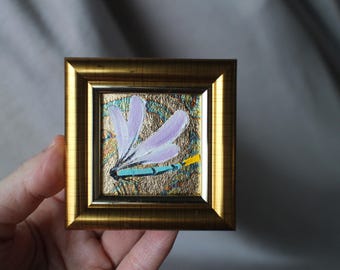 May include: A small framed painting of a blue and purple dragonfly with yellow accents on a gold background.