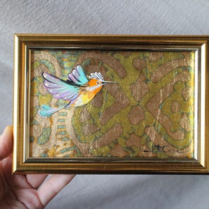 May include: A framed painting of a colorful hummingbird with a blue, purple, and orange body, flying against a gold and green patterned background.