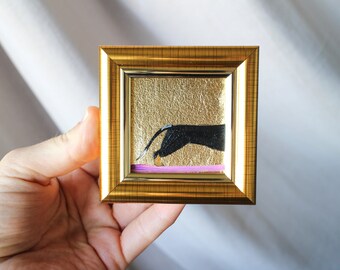 May include: A small framed painting of a black hand reaching for a gold object on a pink background. The background is gold leaf.