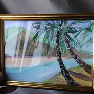 May include: A framed watercolor painting of two palm trees in front of a mountain with a blue and green ocean in the foreground. The painting is in a gold frame.