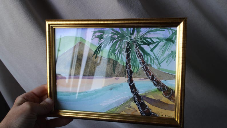 May include: A framed watercolor painting of two palm trees in front of a mountain with a blue and green ocean in the foreground. The painting is in a gold frame.