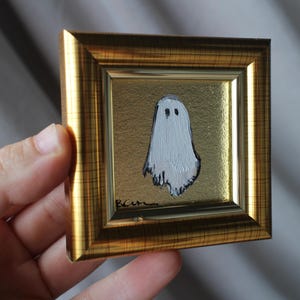 K&ouml;nnte beinhalten: Miniatur-Gem&auml;lde eines wei&szlig;en Geistes mit schwarzen Augen auf goldenem Hintergrund. Das Kunstwerk befindet sich in einem kleinen, quadratischen, goldfarbenen Rahmen. Das Gem&auml;lde hat eine skurrile, handgemachte Qualit&auml;t, perfekt f&uuml;r Halloween-Dekorationen.
