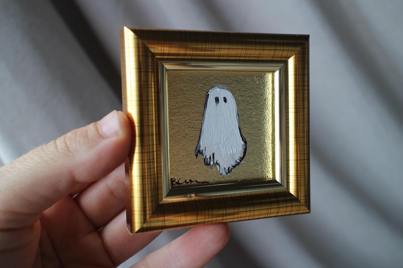 K&ouml;nnte beinhalten: Miniatur-Gem&auml;lde eines wei&szlig;en Geistes mit schwarzen Augen auf goldenem Hintergrund. Das Kunstwerk befindet sich in einem kleinen, quadratischen, goldfarbenen Rahmen. Das Gem&auml;lde hat eine skurrile, handgemachte Qualit&auml;t, perfekt f&uuml;r Halloween-Dekorationen.