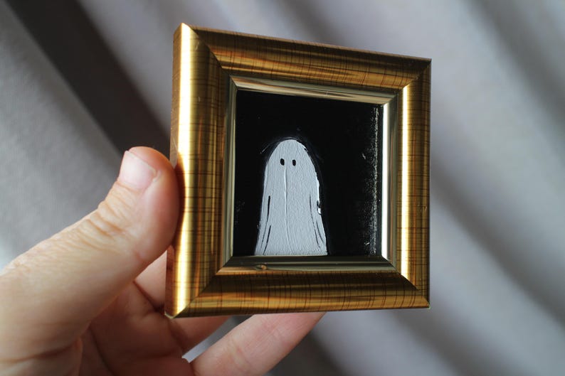 K&ouml;nnte beinhalten: Ein kleines, quadratisches Gem&auml;lde eines wei&szlig;en Geistes mit zwei schwarzen Augen auf schwarzem Hintergrund, gefasst in einem goldfarbenen Rahmen. Das Kunstwerk wird von einer Hand gehalten, was die Miniaturgr&ouml;&szlig;e der gerahmten Kunst zeigt.