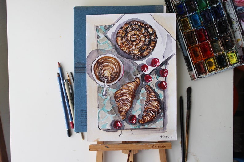 May include: Watercolor painting of a still life featuring a cup of coffee, two croissants, and a plate of cherries. The painting is on a blue background with a white border.