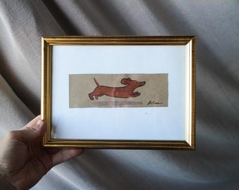 May include: A framed artwork featuring a brown dachshund dog in mid-stride. The dog is depicted with simple lines and a reddish-brown hue against a tan background. The frame is gold-colored.