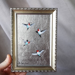 Op de afbeelding: Een ingelijst schilderij van vier kolibries in vlucht tegen een zilveren achtergrond. De kolibries zijn geschilderd in tinten blauw, rood en wit.