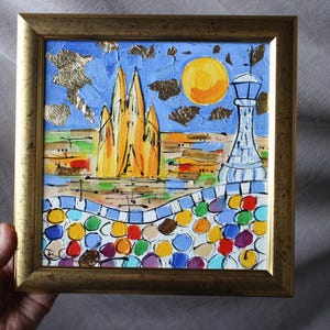 以下が含まれることがあります： 建物、塔、太陽が描かれたカラフルな抽象画。建物は黄色とオレンジ色で、塔は白と青色。太陽は黄色とオレンジ色。背景は青と緑色。絵は金色の額縁に入っている。