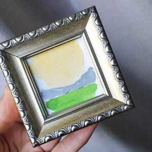 May include: A small framed abstract painting with a yellow, gray, and green color scheme. The painting is in a silver frame with a decorative border.
