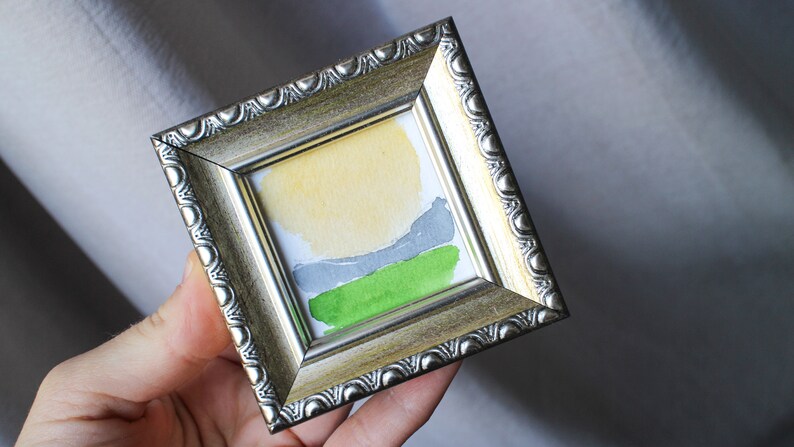May include: A small framed abstract painting with a yellow, gray, and green color scheme. The painting is in a silver frame with a decorative border.