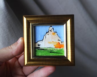 May include: A miniature painting in a gold frame. The artwork depicts a white structure against a blue sky, with green grass and orange accents. The painting is small, likely a few inches in size, and held in a gold-colored frame.