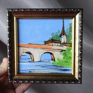 May include: A framed miniature painting depicts a bridge with arches over a river, a church with a steeple, and a blue sky. The painting is in a gold-colored ornate frame.