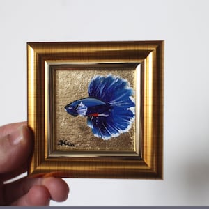 May include: A framed painting of a blue betta fish with a red and white stripe on its body. The fish is swimming with its fins spread out. The painting is on a gold background.