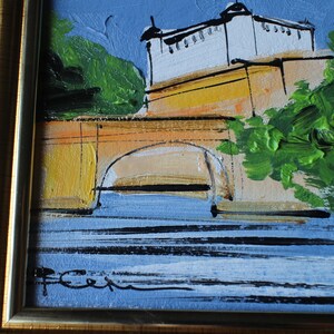 A framed oil painting depicting a building with a white facade and a yellow bridge over blue water. The artwork features green foliage and a blue sky. The painting is signed in the lower left corner and framed in a gold-colored frame.
