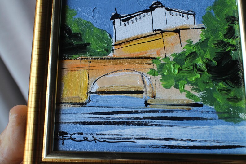 A framed oil painting depicting a building with a white facade and a yellow bridge over blue water. The artwork features green foliage and a blue sky. The painting is signed in the lower left corner and framed in a gold-colored frame.