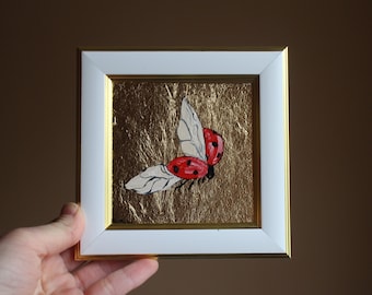 May include: A framed painting of a red ladybug with white wings flying against a gold background.