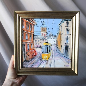 Op de afbeelding: Een ingelijst olieverfschilderij van een stadstaferelen met een gele tram op de voorgrond. De gebouwen zijn geschilderd in een losse, impressionistische stijl met warme kleuren. Het schilderij is onderaan rechts gesigneerd met "KCC".