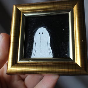 K&ouml;nnte beinhalten: Gerahmtes Gem&auml;lde mit einem wei&szlig;en Geist vor schwarzem Hintergrund. Der Geist hat zwei kleine schwarze Augen. Der Rahmen ist goldfarben mit einer geriffelten Textur. Das Kunstwerk wird in einer Hand gehalten.