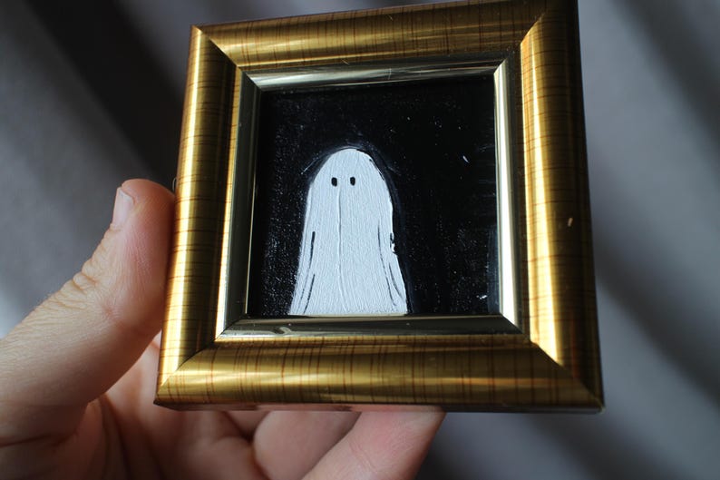 K&ouml;nnte beinhalten: Gerahmtes Gem&auml;lde mit einem wei&szlig;en Geist vor schwarzem Hintergrund. Der Geist hat zwei kleine schwarze Augen. Der Rahmen ist goldfarben mit einer geriffelten Textur. Das Kunstwerk wird in einer Hand gehalten.