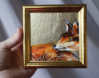 Puede incluir: Una pintura enmarcada de un zorro con pelaje naranja, ojos negros y hocico blanco. El zorro está pintado sobre un fondo dorado.