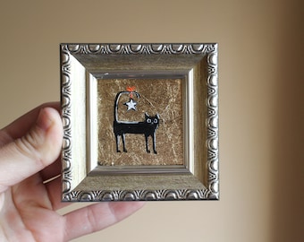 May include: A framed painting of a black cat with a star on its tail, set against a gold background.
