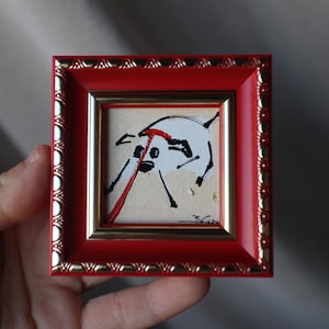 May include: A small, square painting in a red frame with gold trim. The artwork features a white figure with black and red accents. The painting is held in a hand, showcasing the miniature size and artistic style.