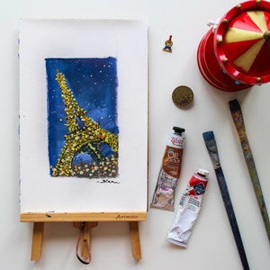 May include: A watercolor painting of the Eiffel Tower in Paris, France. The tower is depicted in a dark blue night sky with twinkling lights. The painting is on a small wooden easel with the word "Artmate" printed on the easel.