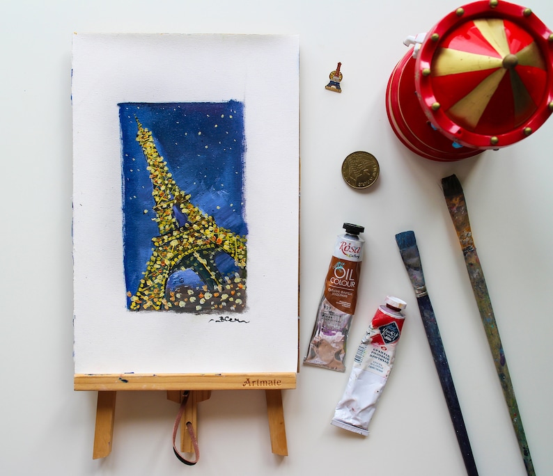 May include: A watercolor painting of the Eiffel Tower in Paris, France. The tower is depicted in a dark blue night sky with twinkling lights. The painting is on a small wooden easel with the word "Artmate" printed on the easel.