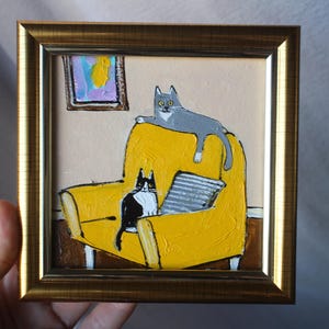 May include: A framed painting of two cats on a yellow armchair. A grey cat reclines on the chair's back, and a black and white cat sits in the seat. A small painting hangs on the wall above the chair.