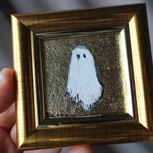 K&ouml;nnte beinhalten: Gerahmtes Gem&auml;lde mit einem wei&szlig;en Geist mit schwarzen Augen auf goldenem, strukturiertem Hintergrund. Das Gem&auml;lde ist in einem goldfarbenen Rahmen gefasst. Das Kunstwerk wird von einer Hand gehalten.