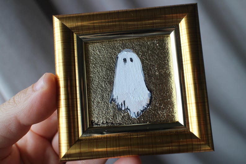 K&ouml;nnte beinhalten: Gerahmtes Gem&auml;lde mit einem wei&szlig;en Geist mit schwarzen Augen auf goldenem, strukturiertem Hintergrund. Das Gem&auml;lde ist in einem goldfarbenen Rahmen gefasst. Das Kunstwerk wird von einer Hand gehalten.