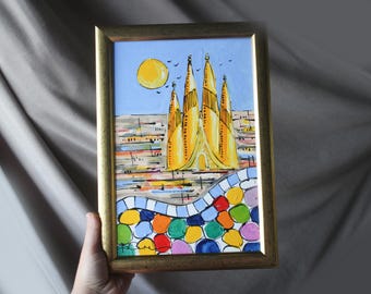 May include: A framed painting depicts a yellow architectural structure under a blue sky with a yellow sun and birds. The artwork features a colorful, mosaic-like pattern at the bottom, with a gold-colored frame. The painting is held by a hand.