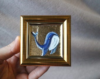 May include: A small framed painting of a blue whale with a white belly, set against a gold background.