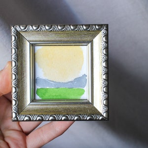 May include: A small framed watercolor painting of a landscape with a yellow sun, gray hills, and green grass.