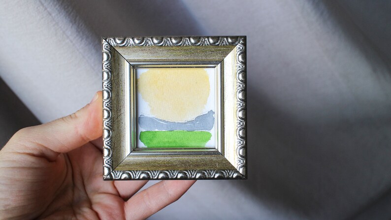 May include: A small framed watercolor painting of a landscape with a yellow sun, gray hills, and green grass.
